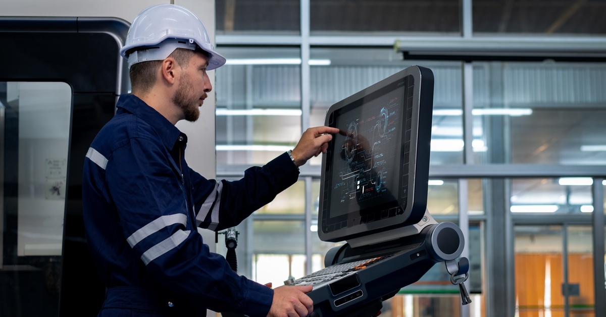 An operator in a work jacket and hard hat pointing to a graph on the screen of a large, sturdy computer.