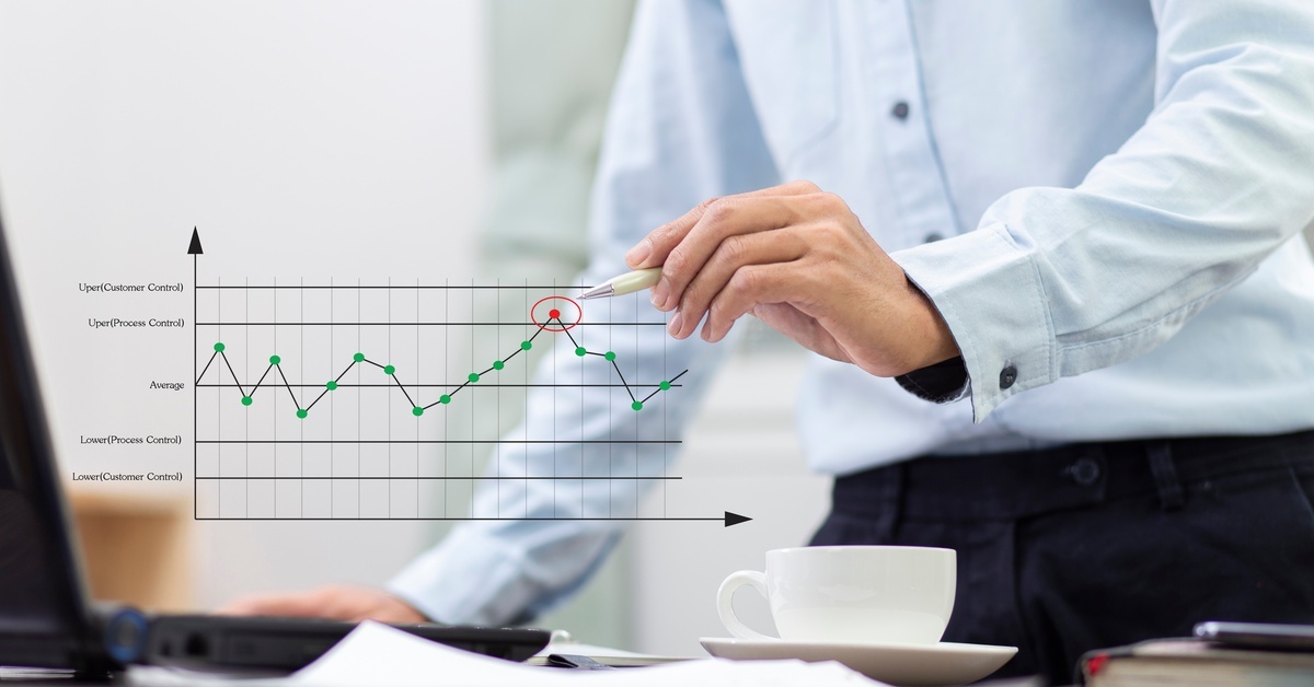 A close-up of a person in business attire pointing a pen toward a red data point on a chart. All other data points are in green.