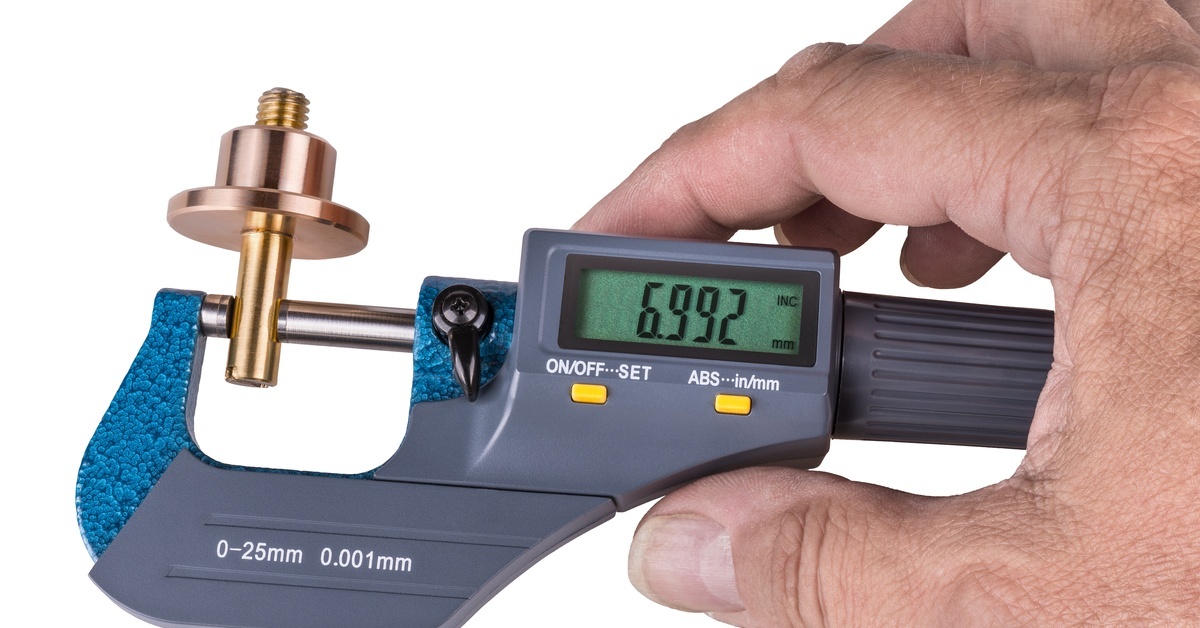 A hand holding a digital micrometer as it measures the screw of a metal piece. The display reads "6.992."
